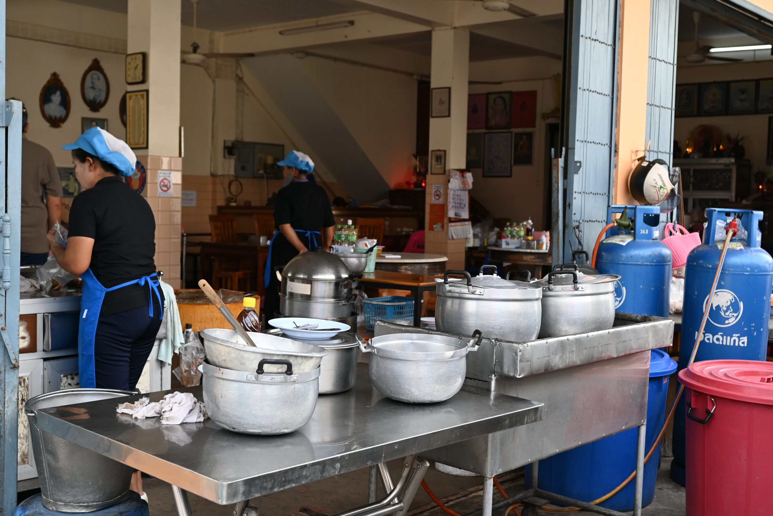 Large aluminum pots lined up on stainless steel prep tables in a busy kitchen with cooks in caps and aprons