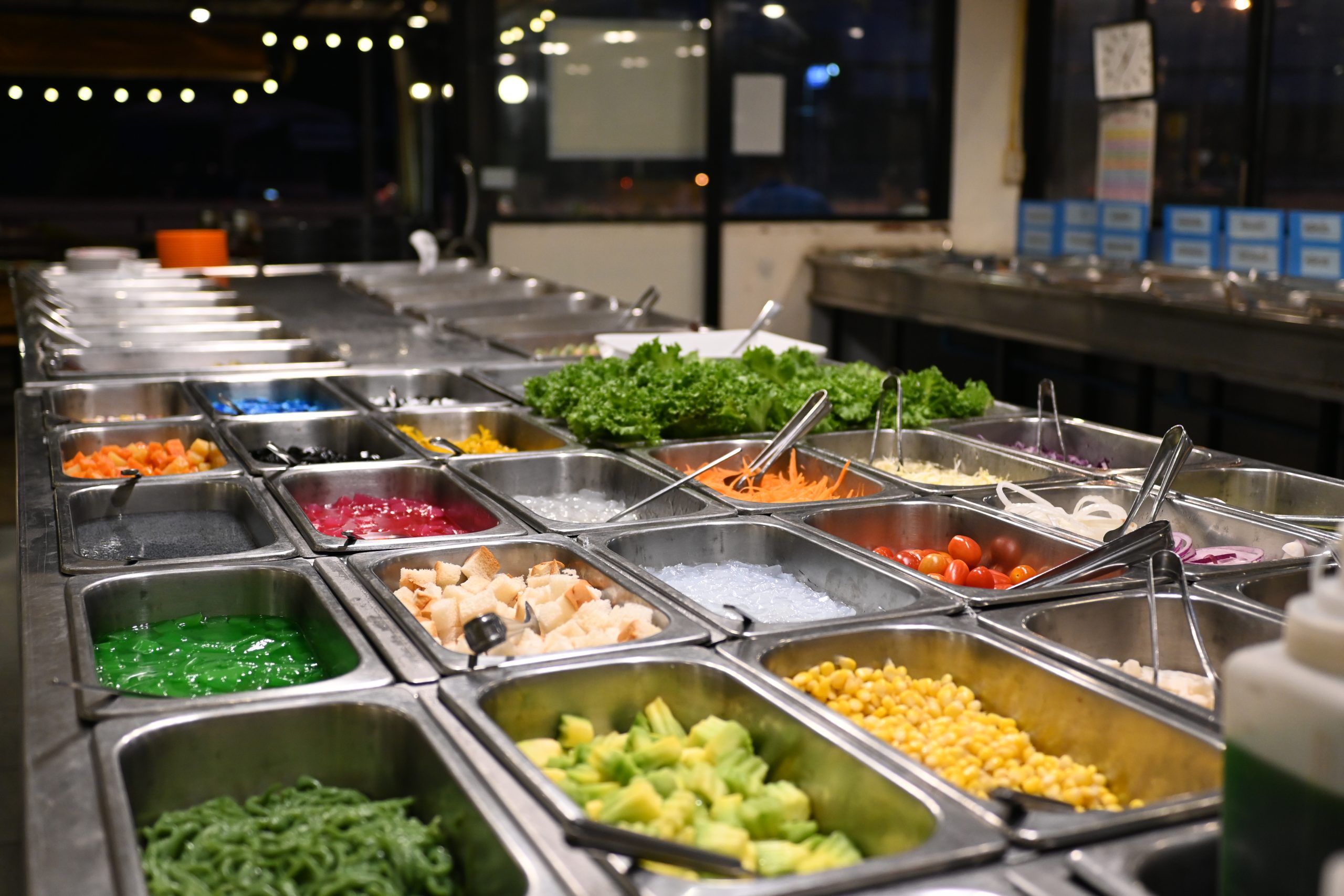 Stainless steel pans of chopped vegetables, greens and toppings held in a refrigerated salad bar