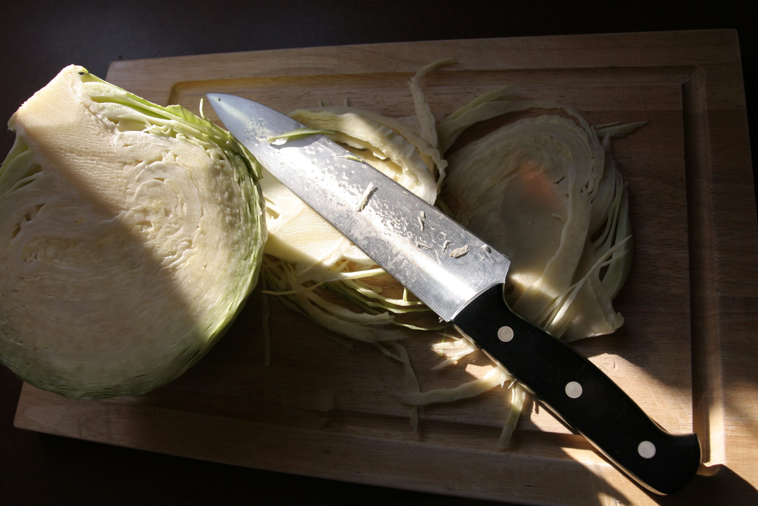 Chef's knife resting on a wooden cutting board beside a halved cabbage and fine shreds