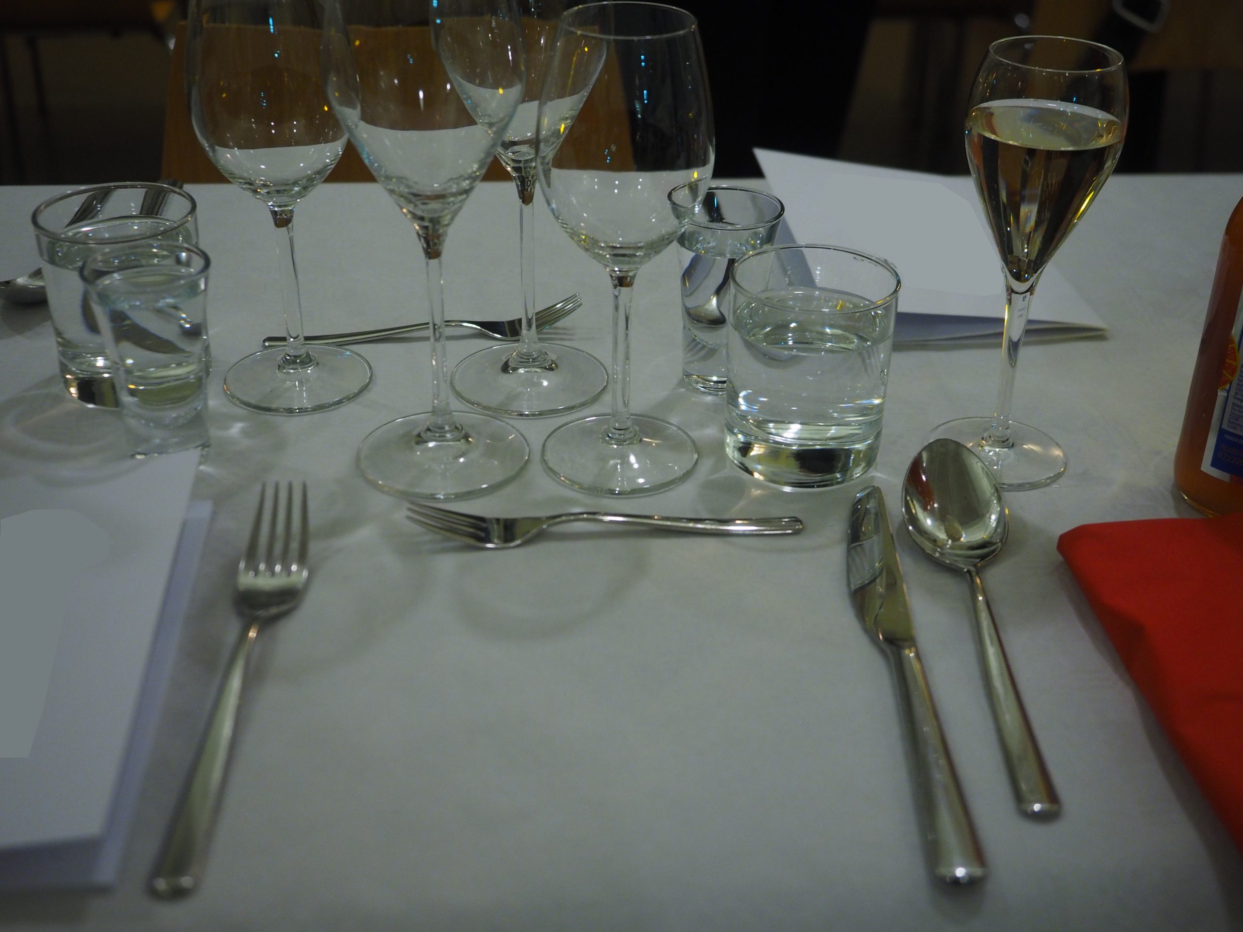 A restaurant place setting with knife, fork, spoon, water tumblers and wine glasses on a white tablecloth