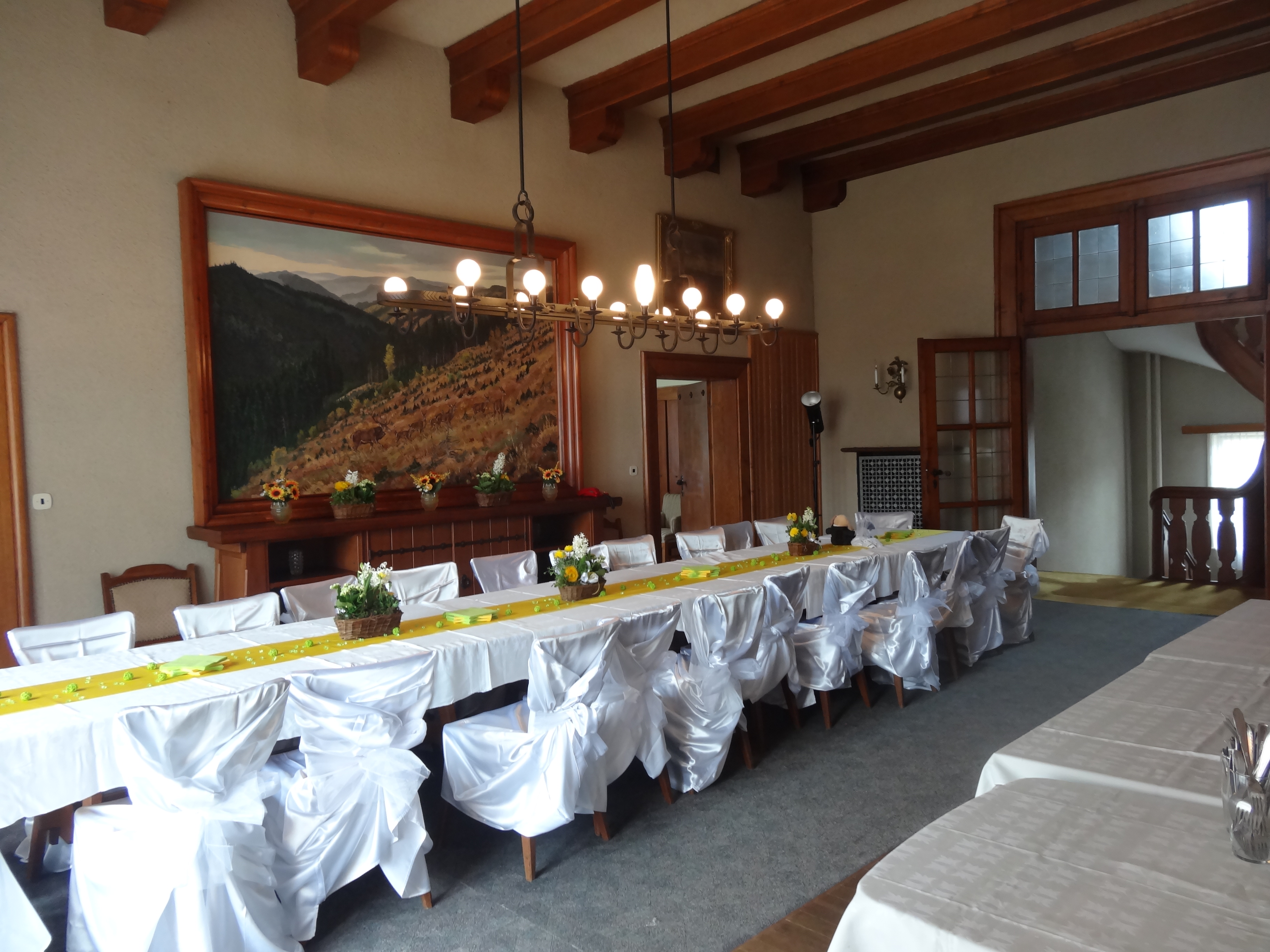 A long banquet table with white chair covers and flowers set up for an event in a wood-beamed hall