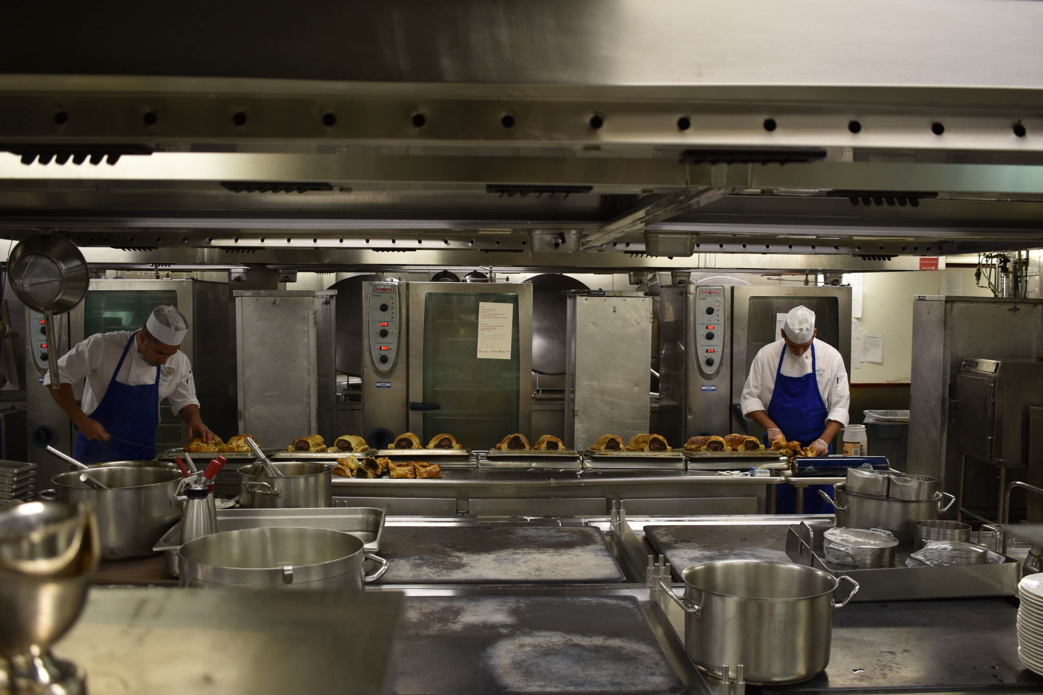 Two cooks in aprons and caps working at stainless steel counters in a large commercial kitchen