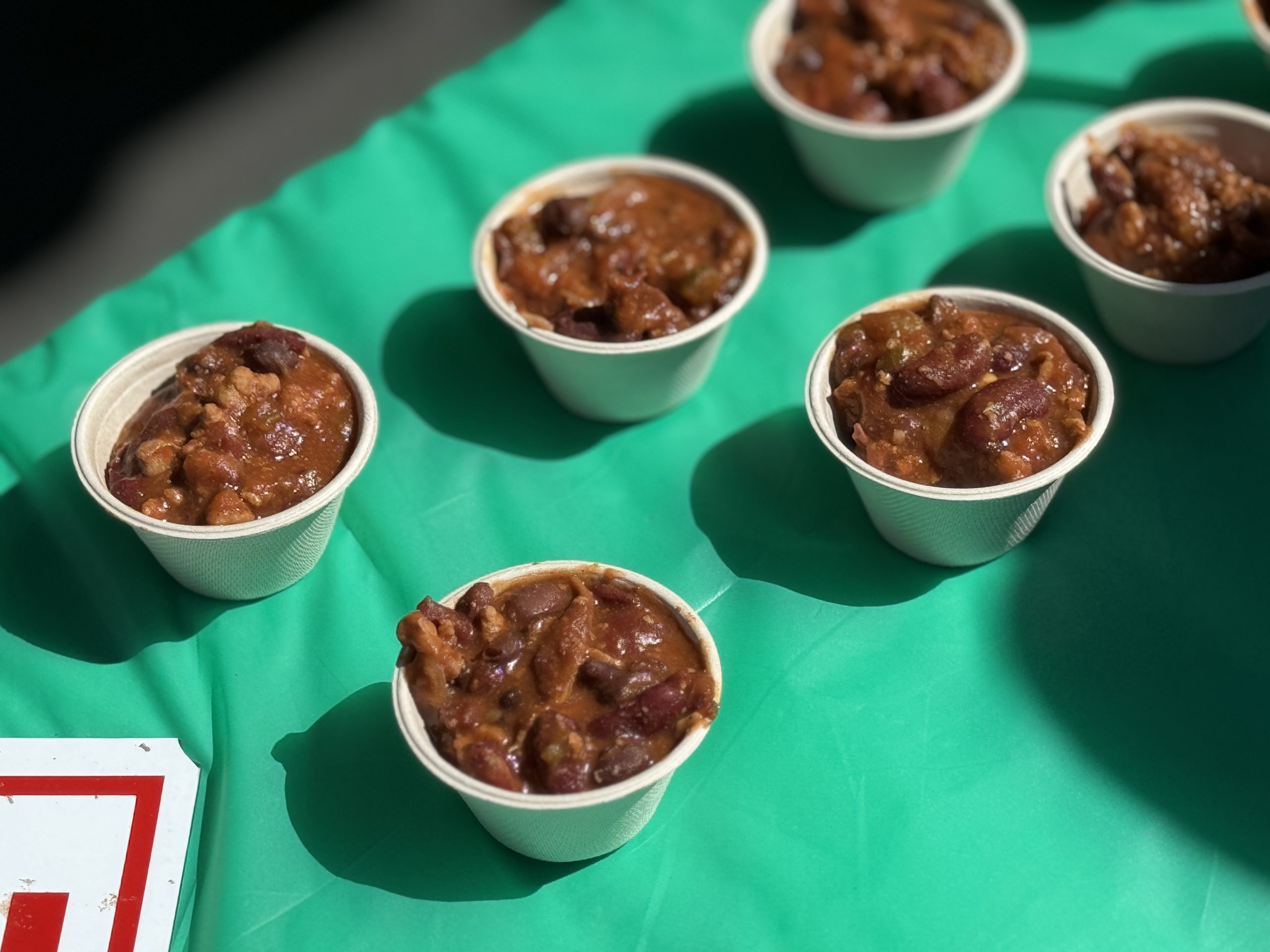 Small paper cups of bean chili lined up on a green tablecloth for tasting