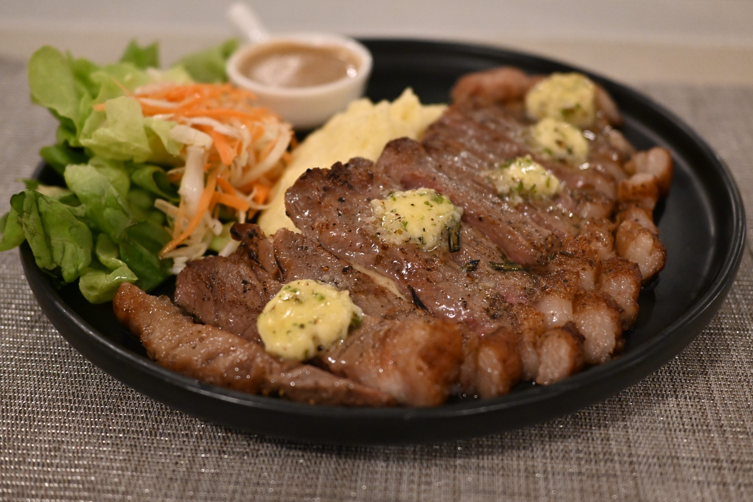 Sliced grilled steak with herb butter, mashed potato, salad greens and a small dish of gravy