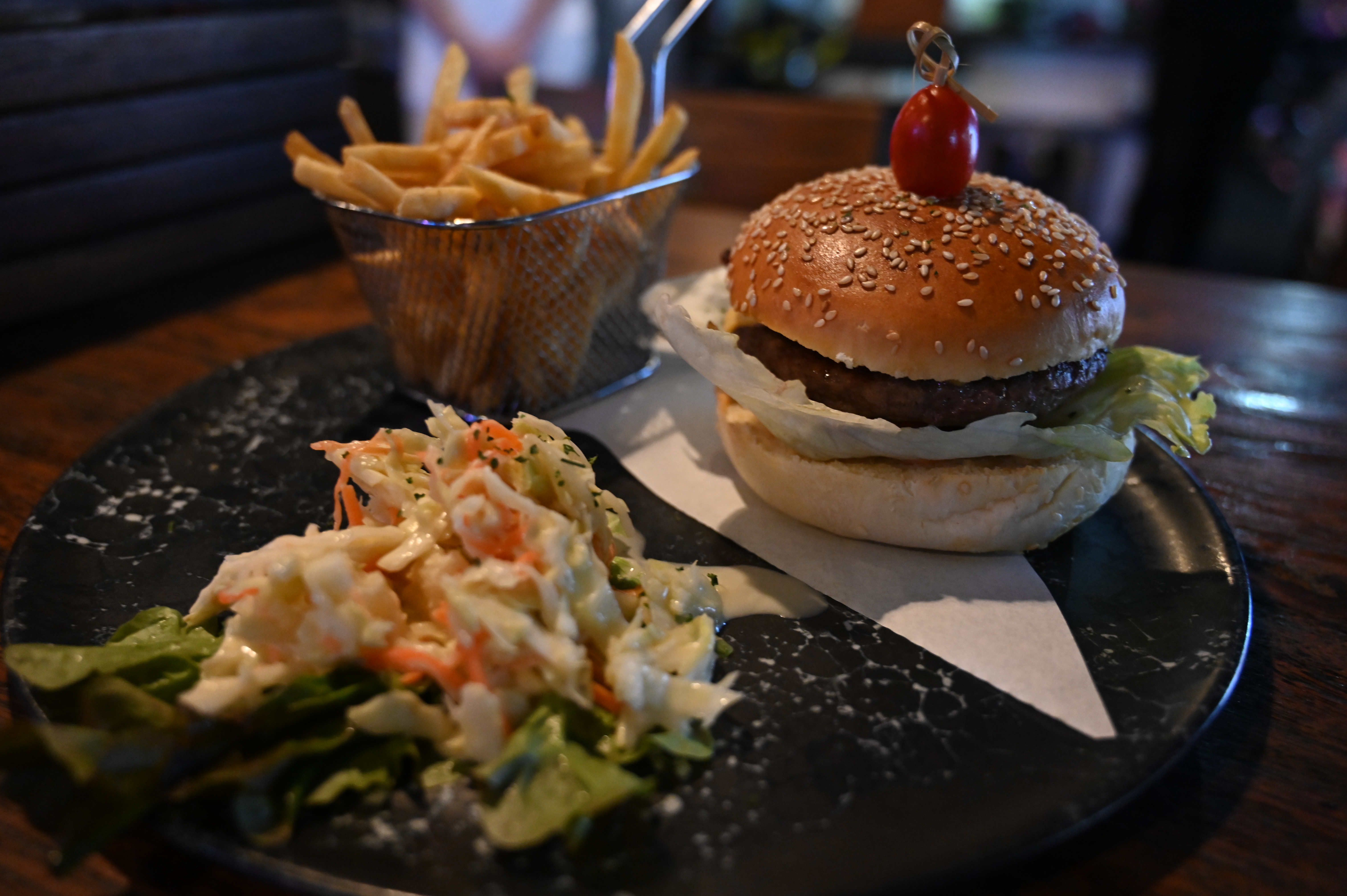 Burger on a sesame bun with a basket of fries and a side of coleslaw on a dark plate