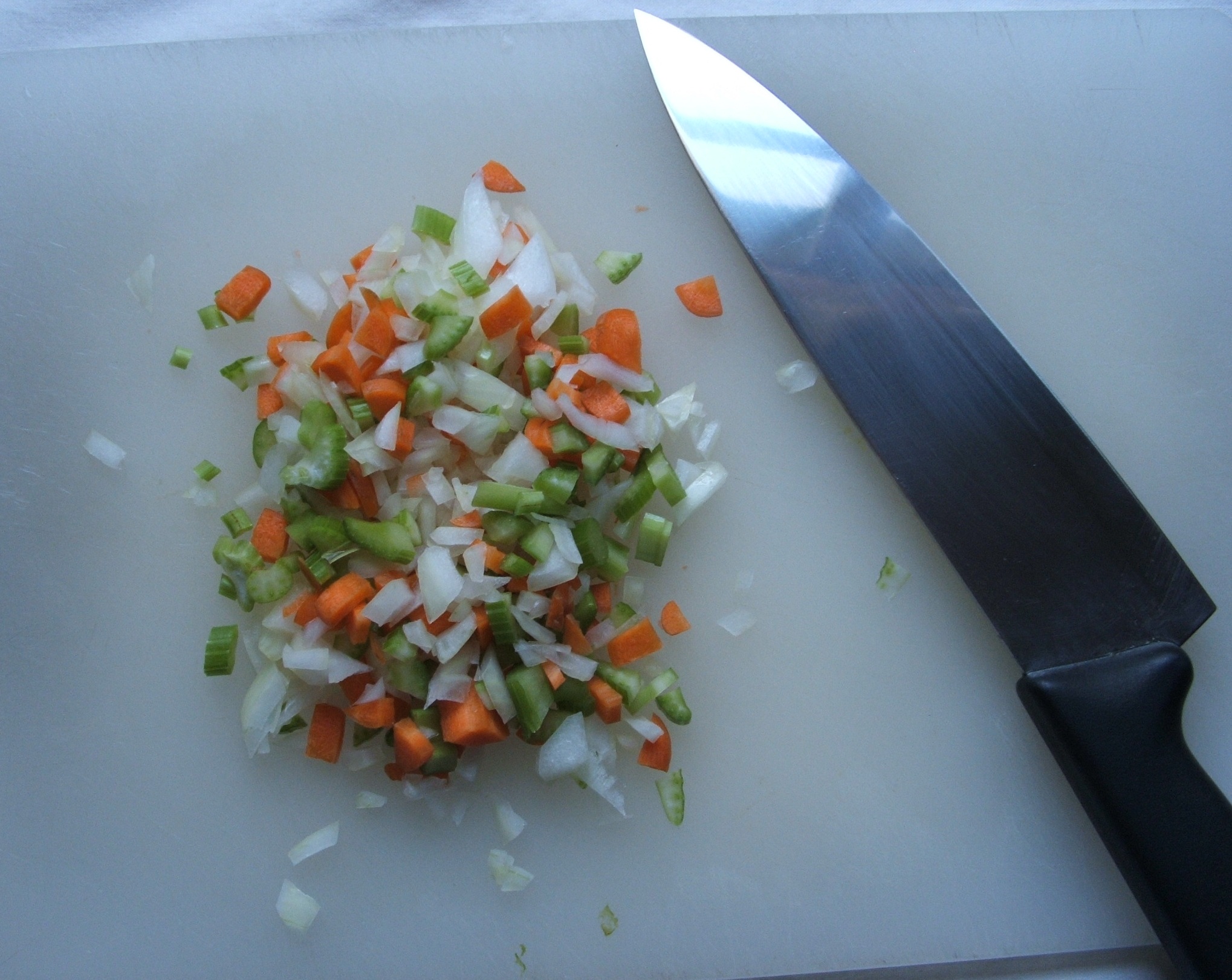 Finely diced carrot, celery and onion on a white cutting board beside a chef's knife